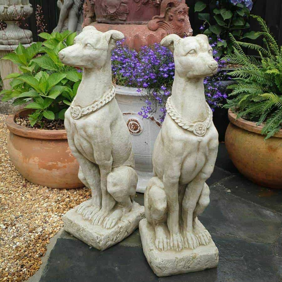 A pair of regal hound statues sitting at attention. Situated on the stone floor of a British garden with potted plants in the background