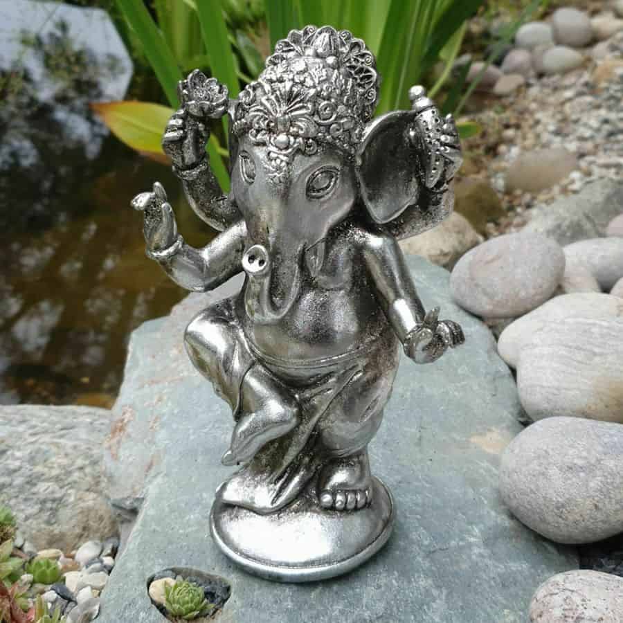 A ganesh statue of silver finish featuring an elephant head and gour arms. Situated amongst the stones of a British garden