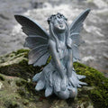 A fairy on her knees with long flowing hair and extended wings of grey finish. Situated on the mossy rock of a British garden
