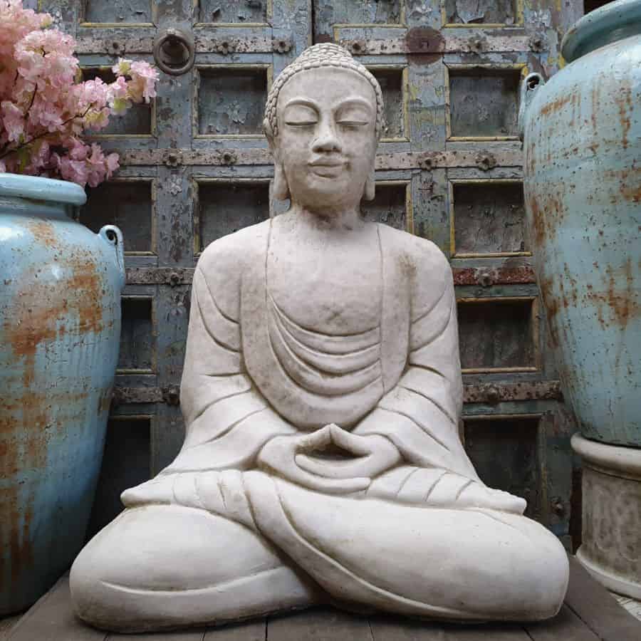 A substantial buddha with flowing robes, peaceful expression and legs crossed in a meditation position. Situated in the garden of a British home with blue pots in background