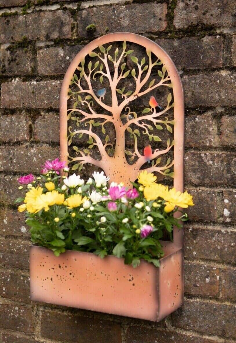 A metal sheet with planter at the base filled with yellow and pinks flowers. Adorned with the tree of life with green leaves and little birds. Hanging on the wall of a British garden