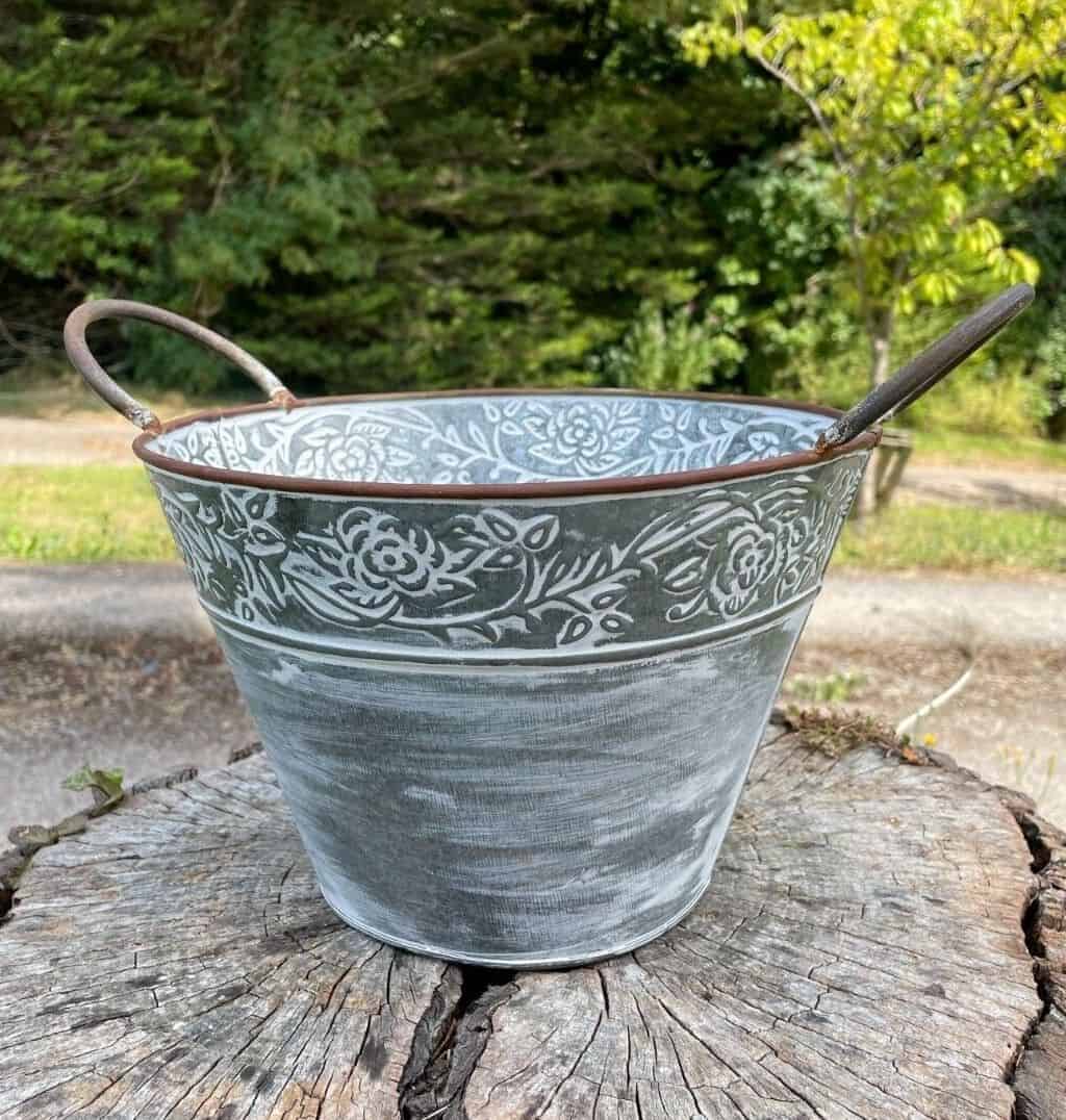 A plant pot of tin with flower decoration inlay around the rim. Two handles of brown finish adorning the top of the pot. Situated on a treestump in a British garden