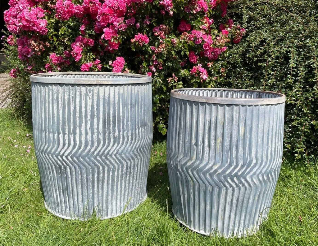 Two dolly tubs of steel finish with chevron pattern. Situated on the grass of a British garden with pink flowers in the background