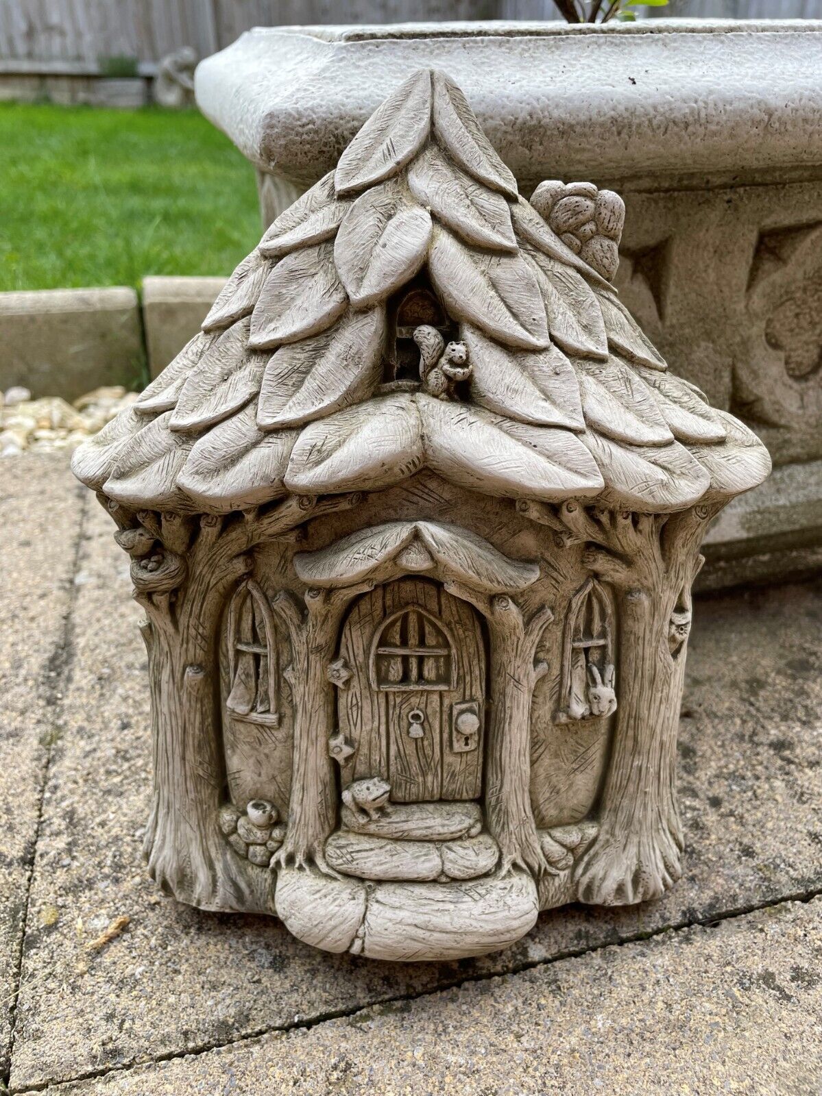 A stone Fairy house feauturing leaves for a roof, squirrel and oak archways framing the front door. Situated in a UK Garden infront of a flower pot and green grass