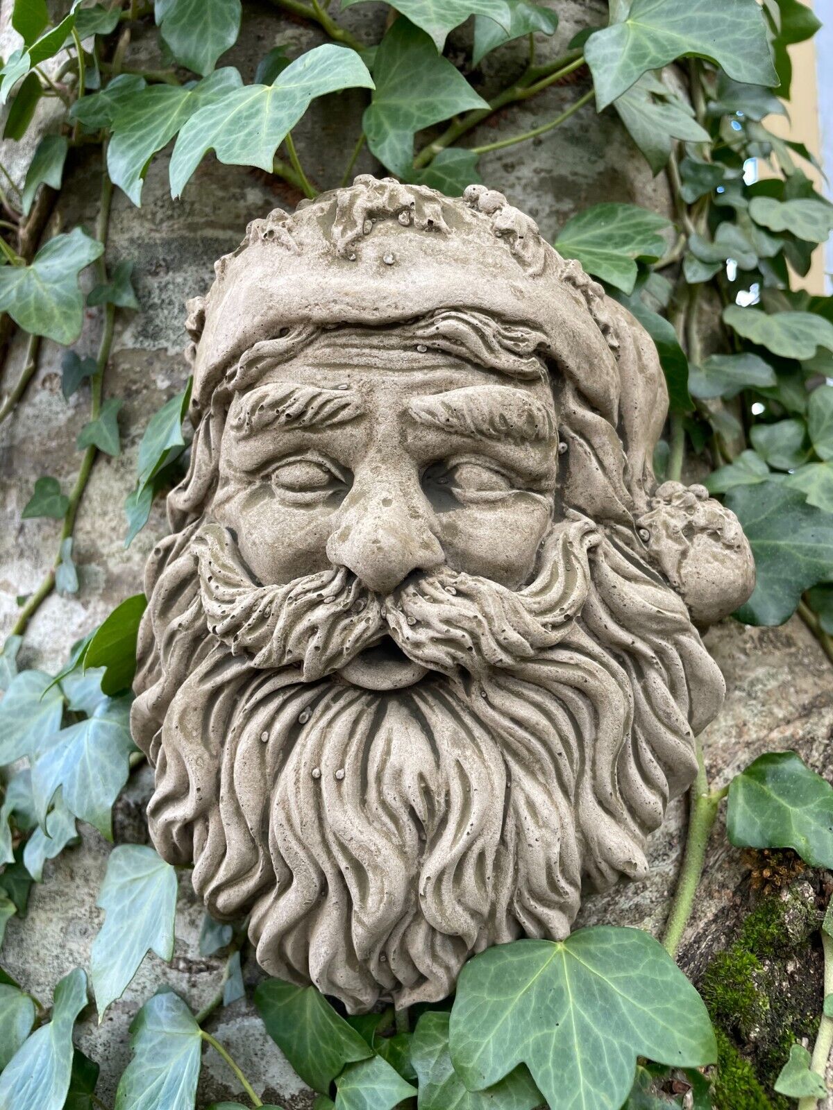 Santa's face in stone statue form, with classic long flowing beard and warm hat with bauble. Situated amongst the ivy in a British Garden