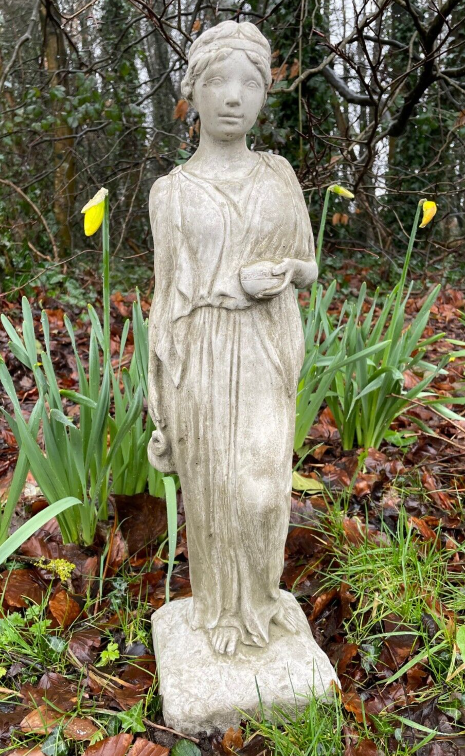 A lady with flowing robes and tied back hair, standing amongst the sprouting yellow daffodils in a British garden