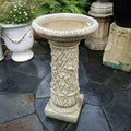 A wide-stemmed birdbath decorated with intricate ivy from top to bottom. Situated on the patio of a British garden