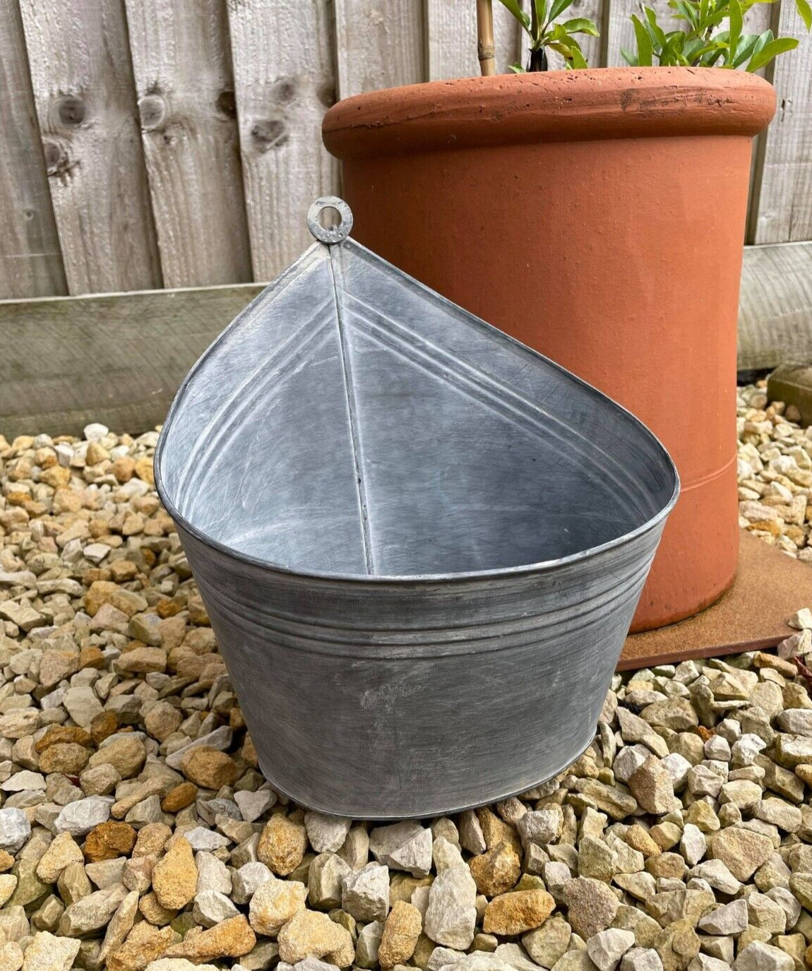 An oval hanging basket of silver coloured zinc with loop for hanging. situated in the gravel of a british garden