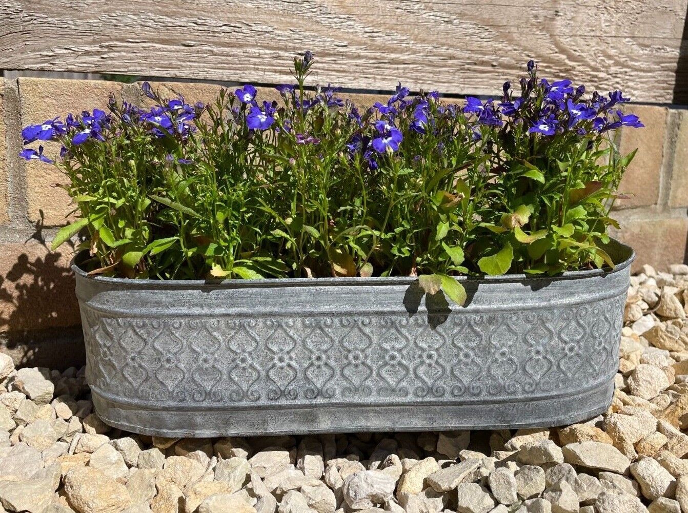 A floral patterned planter of oval shape and silver metal zinc colour featuring planted blue flowers. Situated on the gravel of a British garden