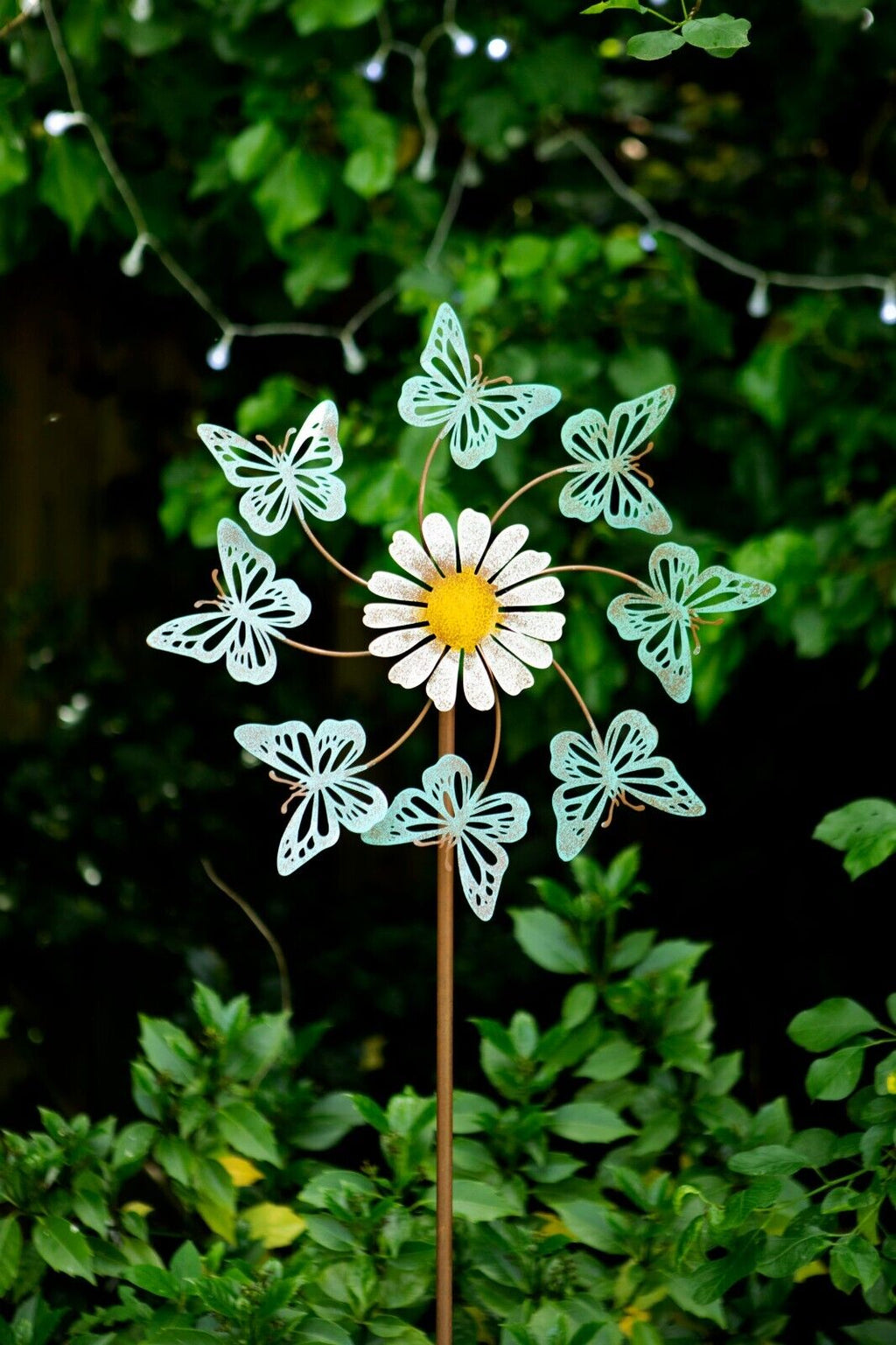 A lovely daisy with 8 butterflies surrounding it on a spinning mechanism. Sticking into the ground of a british garden with foliage in the background