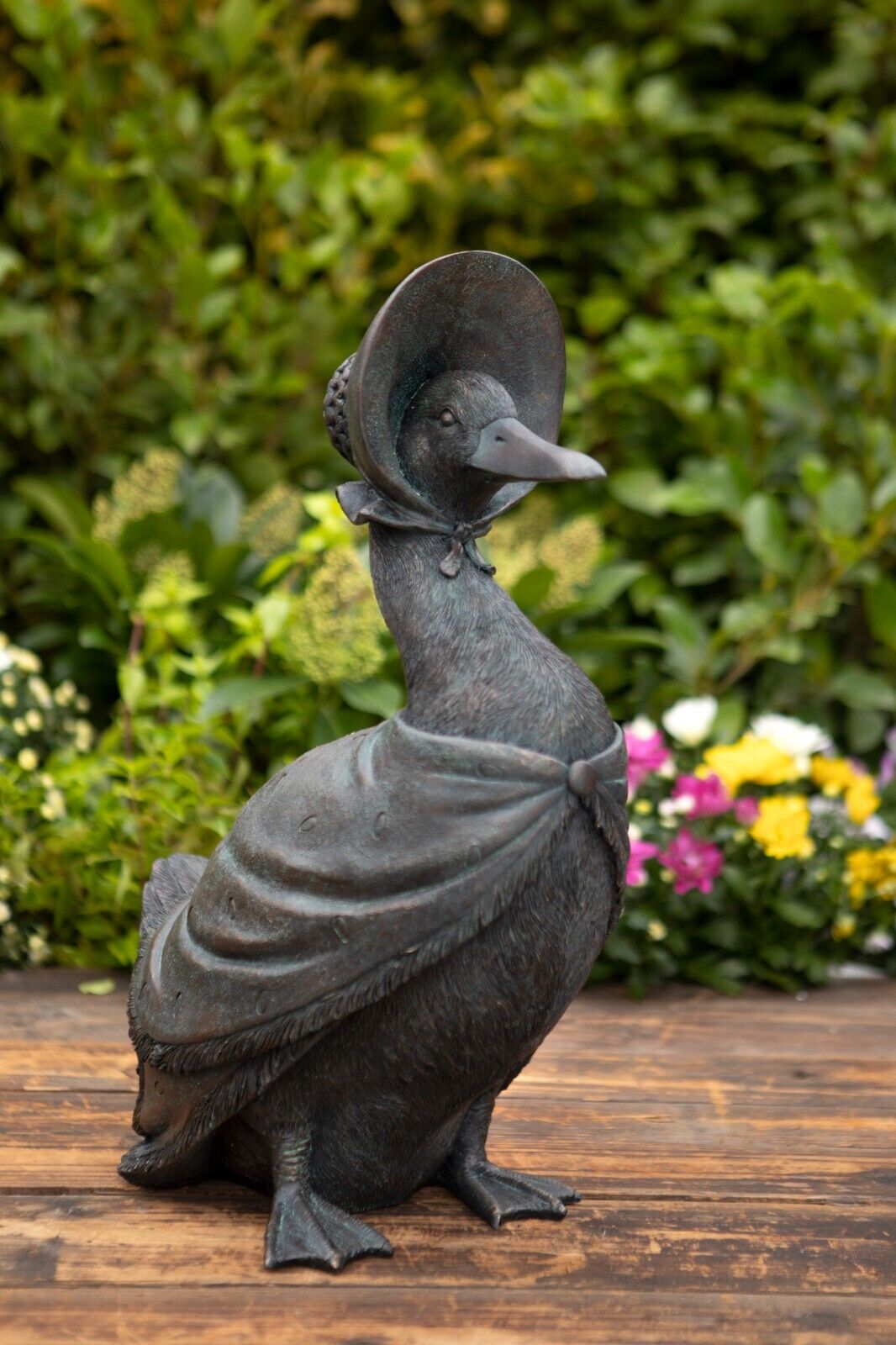 A dark bronze finished Jemima puddle duck with travel cloack and headwear. Situated on the decking of a British garden with flowerbeds and foliage in the background.