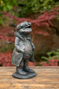 A Mr Toad sculpture adorned with trench coat, bow tie and driving hat/glasses. Situated in a British garden with foliage in the background