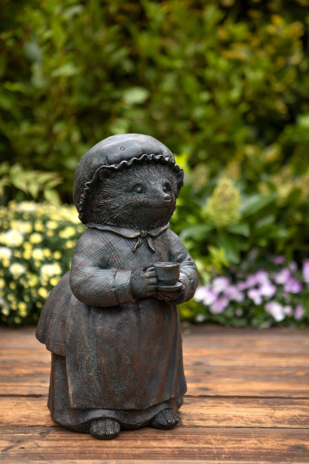 A anthropomorphised hedgehog statue featuring nightcap and cup of tea. Situated on the decking of a british garden with colourful flora in the background