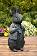 A peter rabbit featuring carrot and waistcoat of dark finish. Situated on the decking of a British garden with colourful flora in the background
