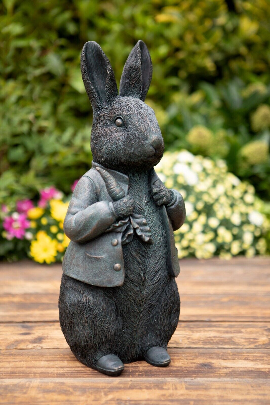 A peter rabbit featuring carrot and waistcoat of dark finish. Situated on the decking of a British garden with colourful flora in the background