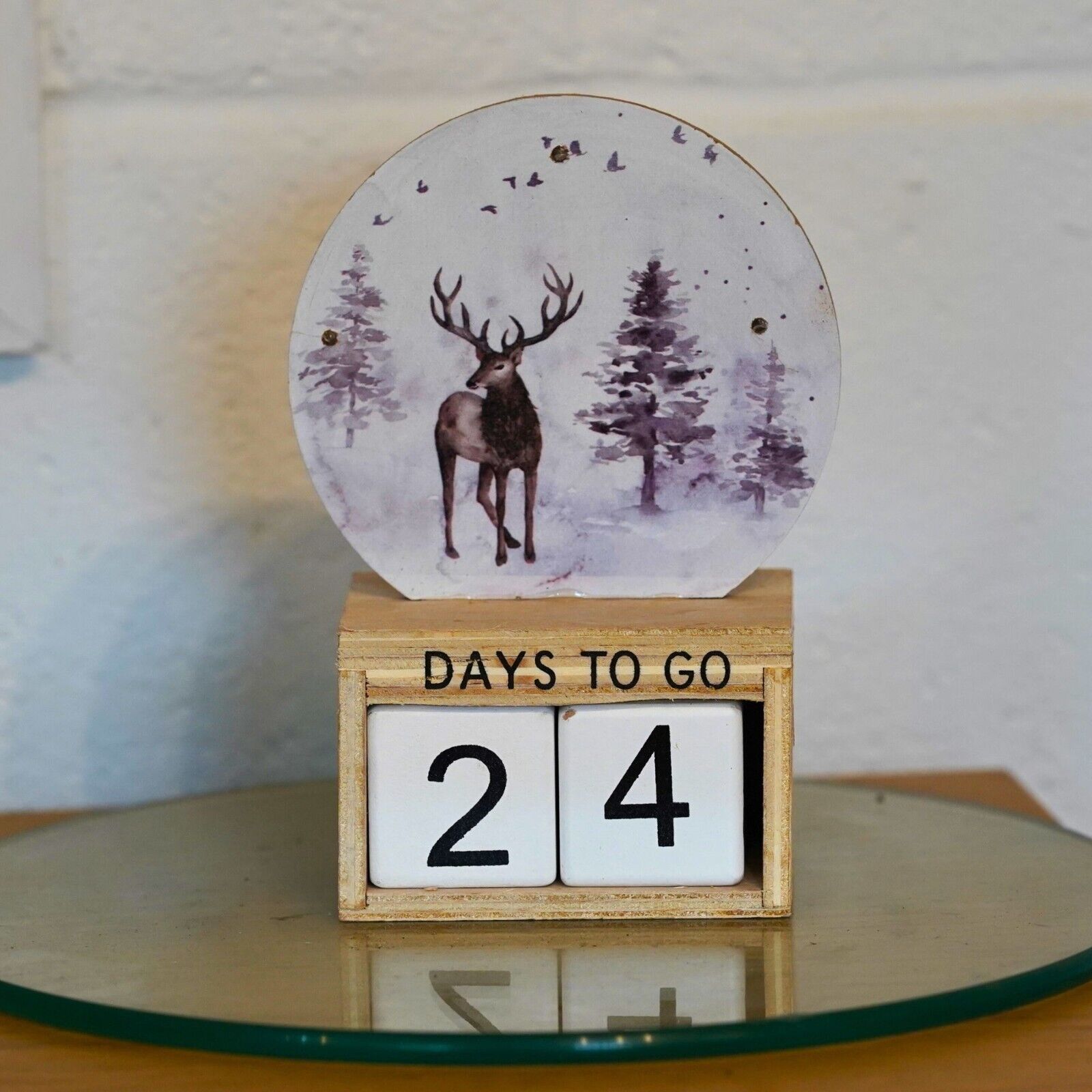 A painted stag reindeer on a china plate, placed on it's side within a wooden advent calendar reading: "Days to go 24". Situated on a glass plinth in a British home