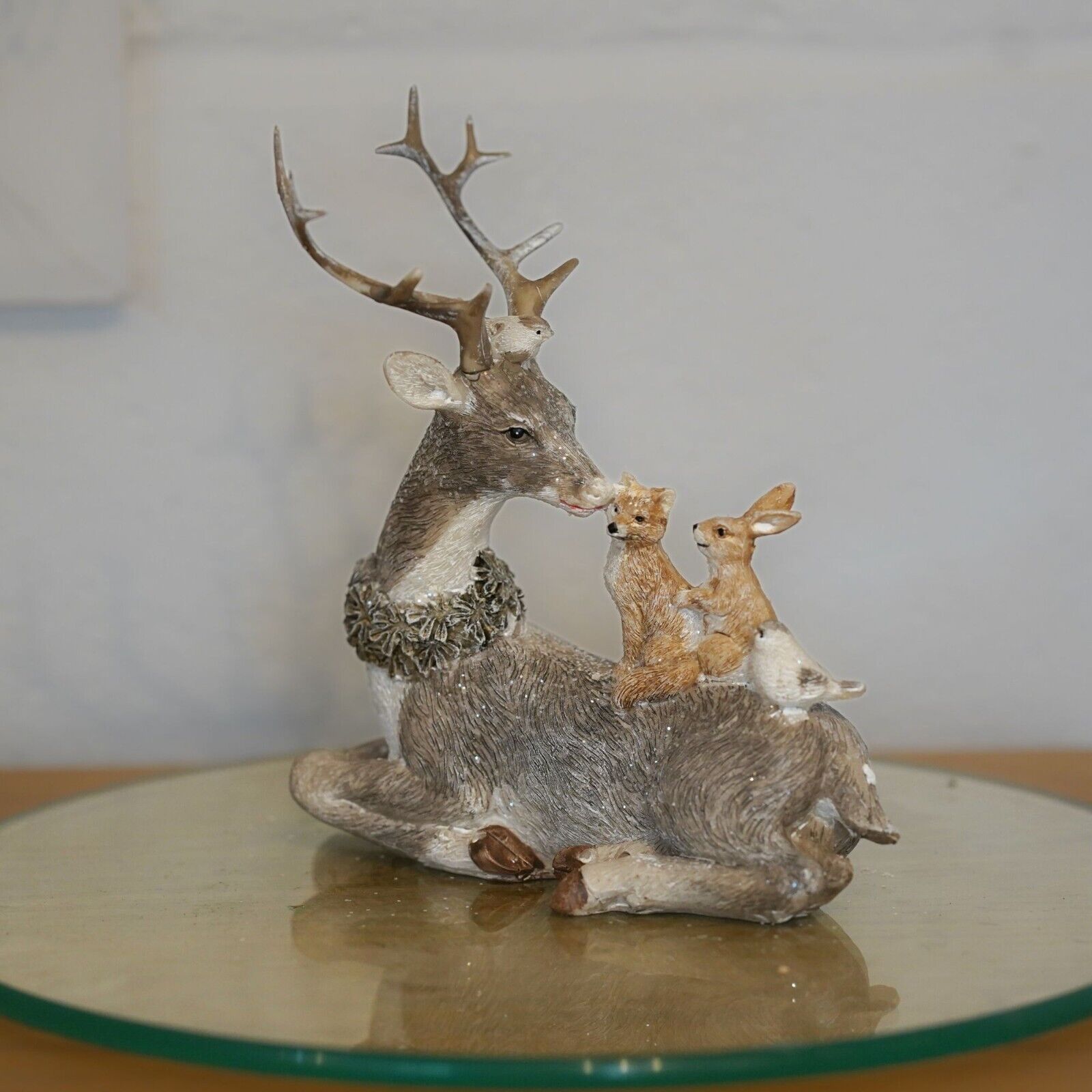A grey reindeer with antlers and wreath around neck, laying with forest critters on back. Situated on a glass plinth in a British home