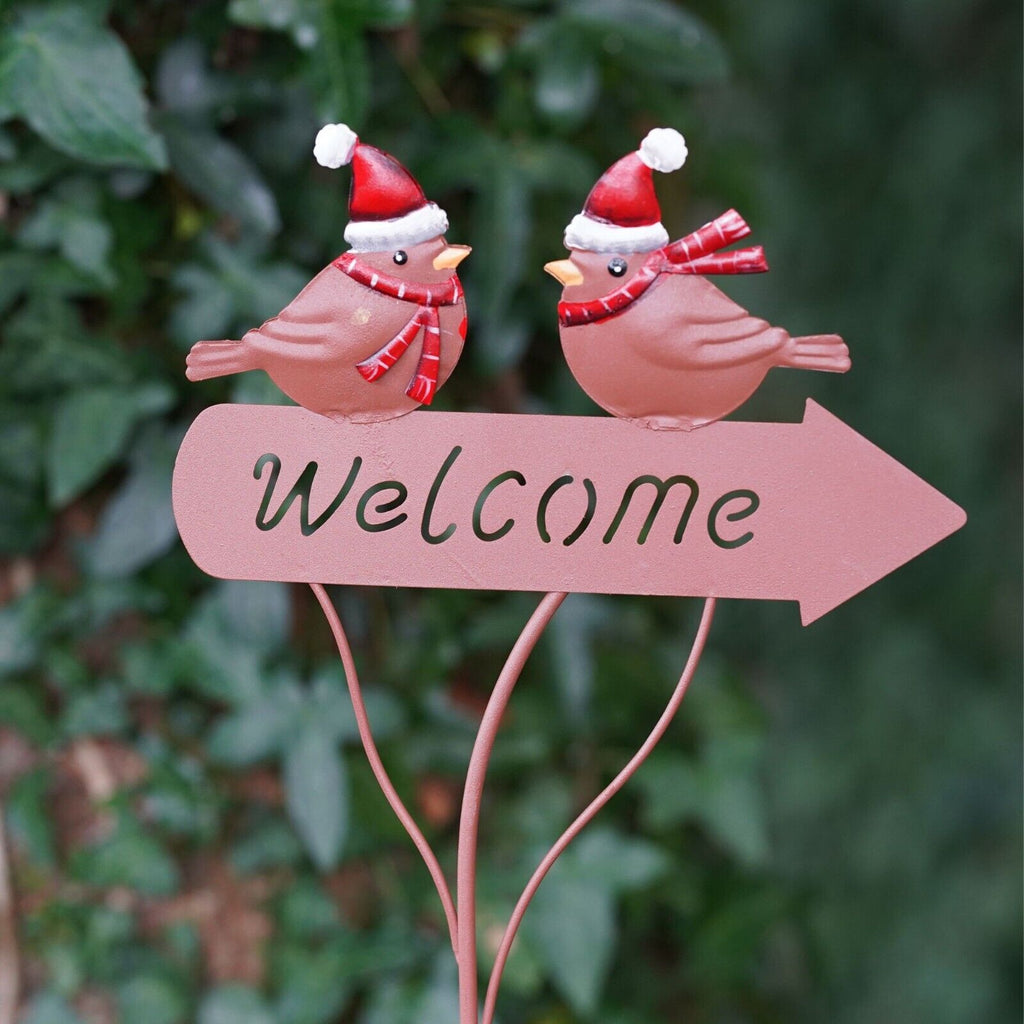 two little robins with red Christmas hats and scarfs, perched upon a welcome sign in the shape of an arrow. All in copper colour. Situated amongst the foliage of a British garden