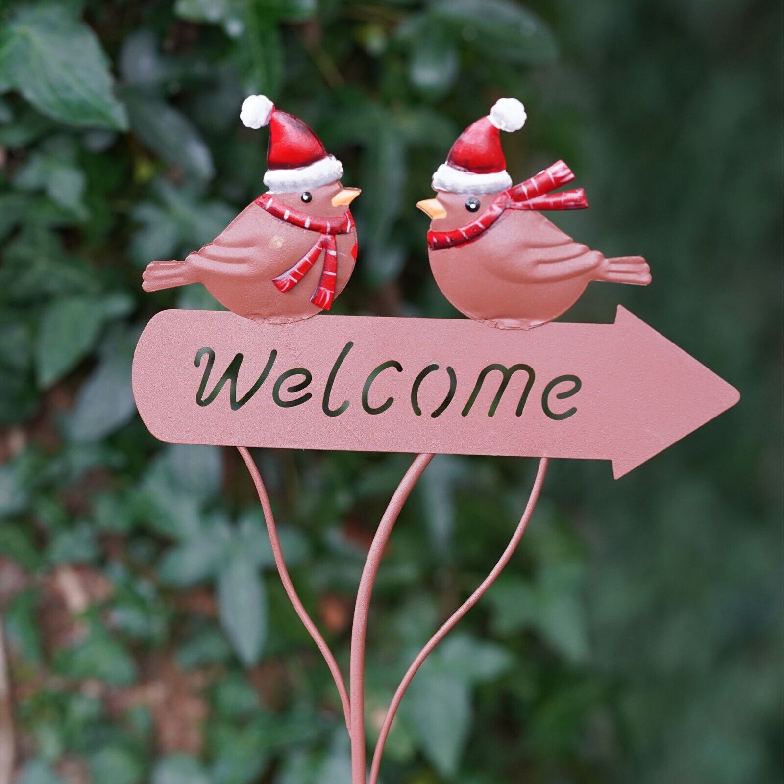 two little robins with red Christmas hats and scarfs, perched upon a welcome sign in the shape of an arrow. All in copper colour. Situated amongst the foliage of a British garden