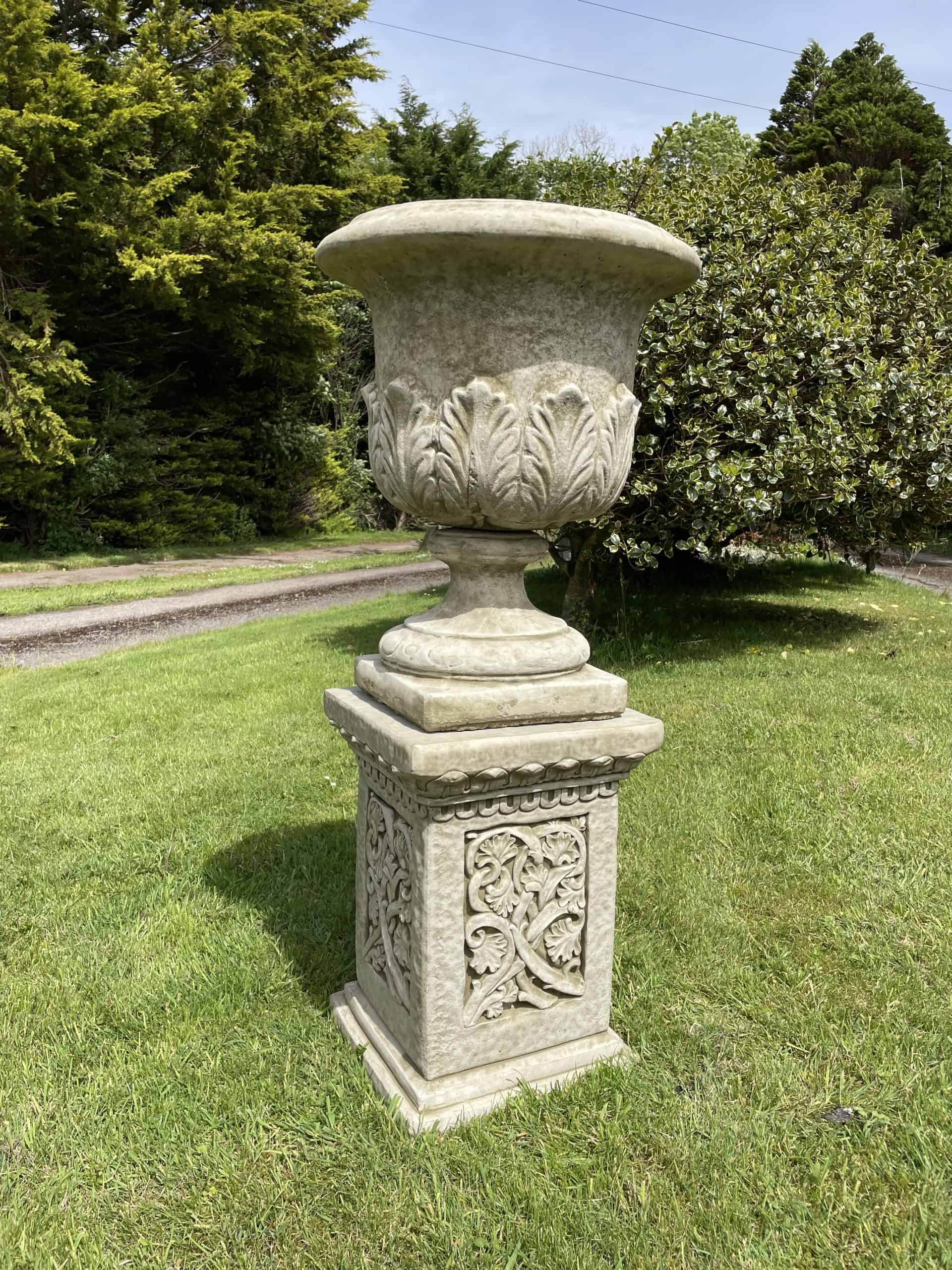A stone urn with leaf detailing and large stone plinth with intricate carvings on the sides. Situated amongst the grass of a British garden