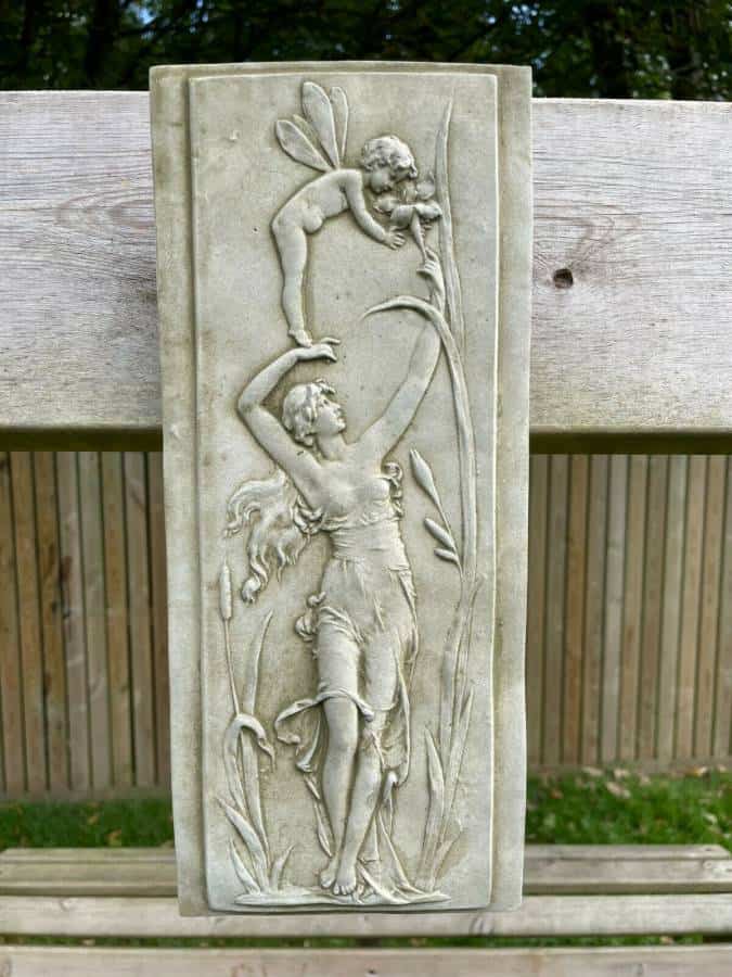 A rectangular stone plaque adorned with a cherub balanced on a nymph's outstretched arm. Situated on a british garden's fence.