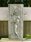 A rectangular stone plaque featuring a cherub balancing on a nymphs hand, feeding them grapes from the vine. Situated on a british garden fence.