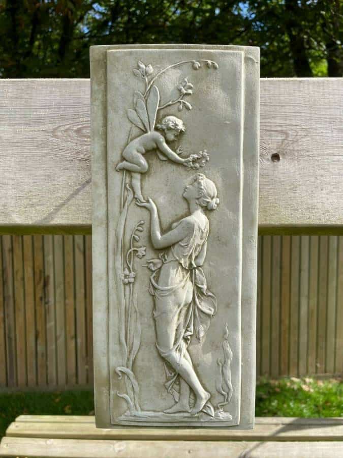 A rectangular stone plaque featuring a cherub balancing on a nymphs hand, feeding them grapes from the vine. Situated on a british garden fence.