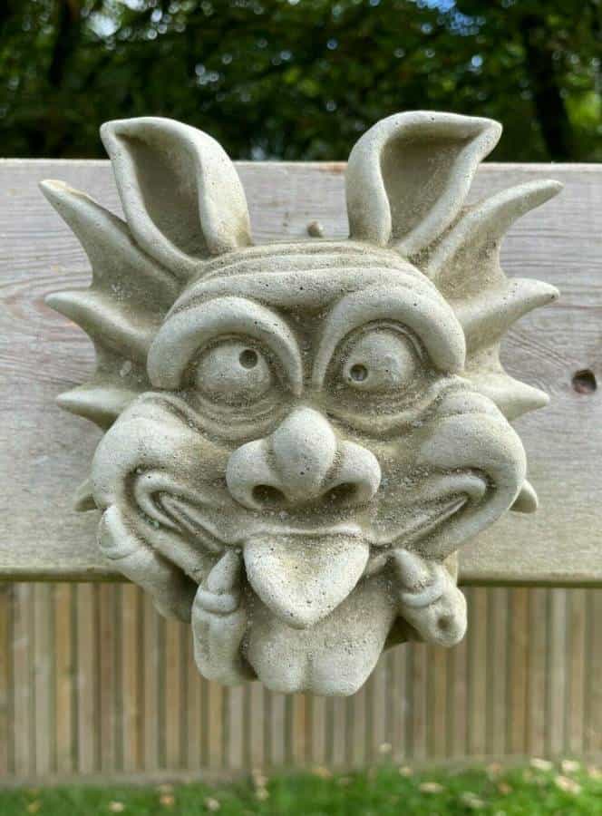 A gremlin face with tongue poked out and eyes crossed cheekily, hanging on a fence in a british garden