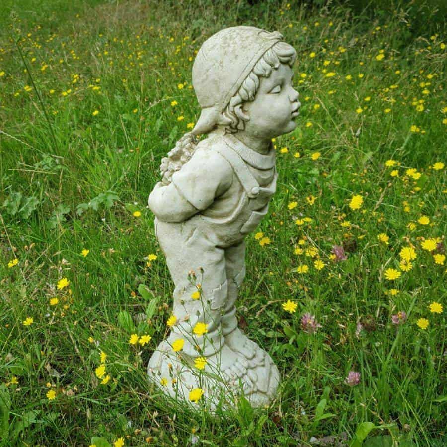 A boy with curly hair and backwards cap with hands behind their back holding flowers. Lips pursed as if to kiss. Situated amongst the green grass of a British garden with yellow daisies