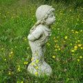 A boy with curly hair and backwards cap with hands behind their back holding flowers. Lips pursed as if to kiss. Situated amongst the green grass of a British garden with yellow daisies