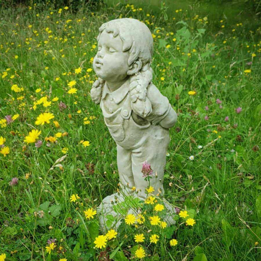 A young girl with pig tails and dungarees. Standing with hands behind her back and lips pursed as if to kiss. Situated amongst the green grass of a British garden with yellow daisies