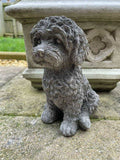 A sitting labradoodle puppy with detailed, fluffy fur made of stone. Situated on the stone flooring of a British garden