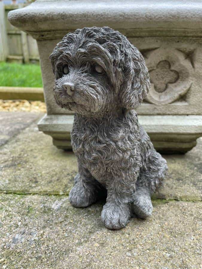 A sitting labradoodle puppy with detailed, fluffy fur made of stone. Situated on the stone flooring of a British garden