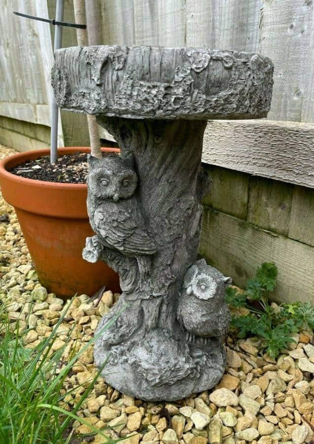 A Rustic birdbath of stone featuring two life-like owls carved into the base. Situated in front of a garden fence in a British garden