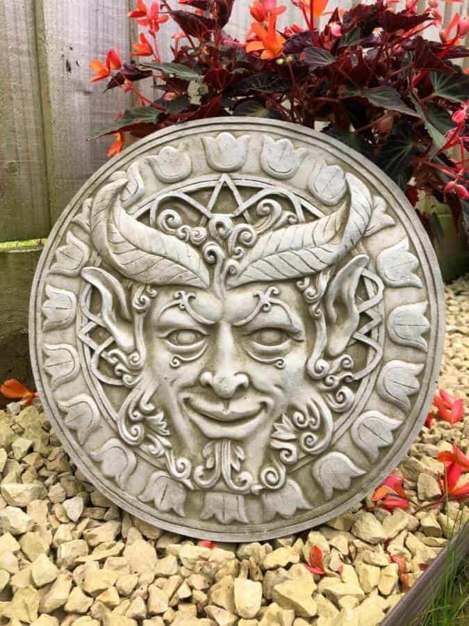 A Greenman from folklore with mysterious eyes carved into a circular plaque featuring intricate patterns and a floral design on the circumference. Situated amongst orange flowers in a british garden