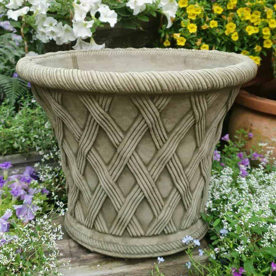 A large urn with intricate criss-cross pattern. Situated on the wooden decking of a British garden, surrounded by colourful flowers.