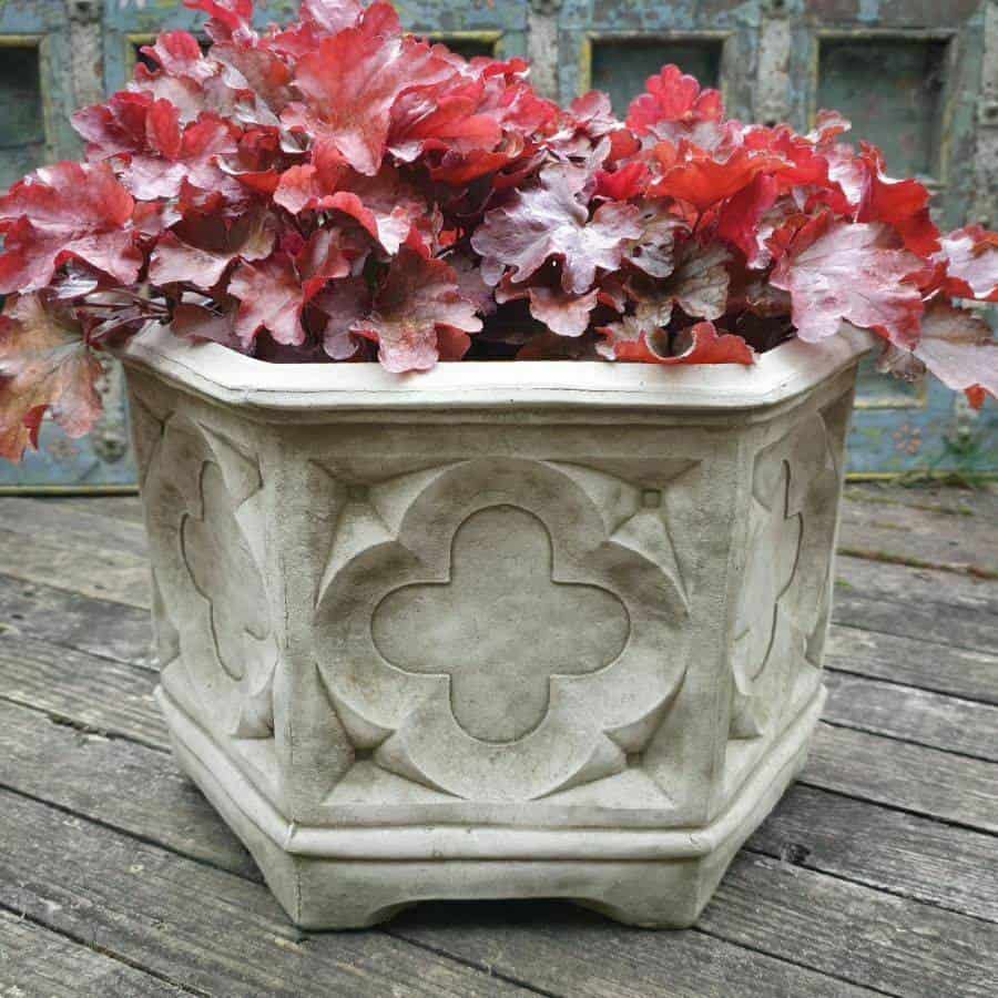 A stone planter with celtic detailing pattern featuring red plants. Situated on the decking of a British garden