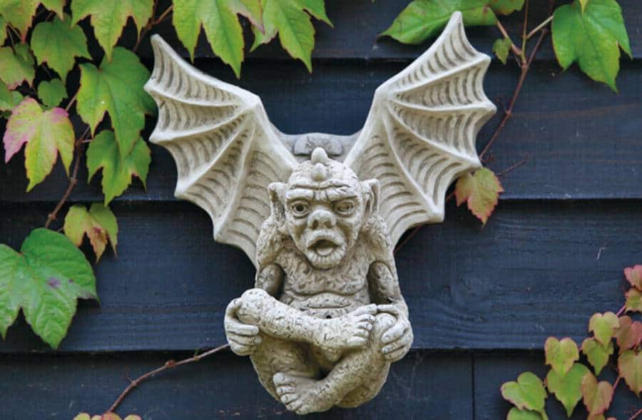 A gargoyle with impressive splayed wings and folded legs hanging on the dark fence of a British garden with green leaves surrounding them