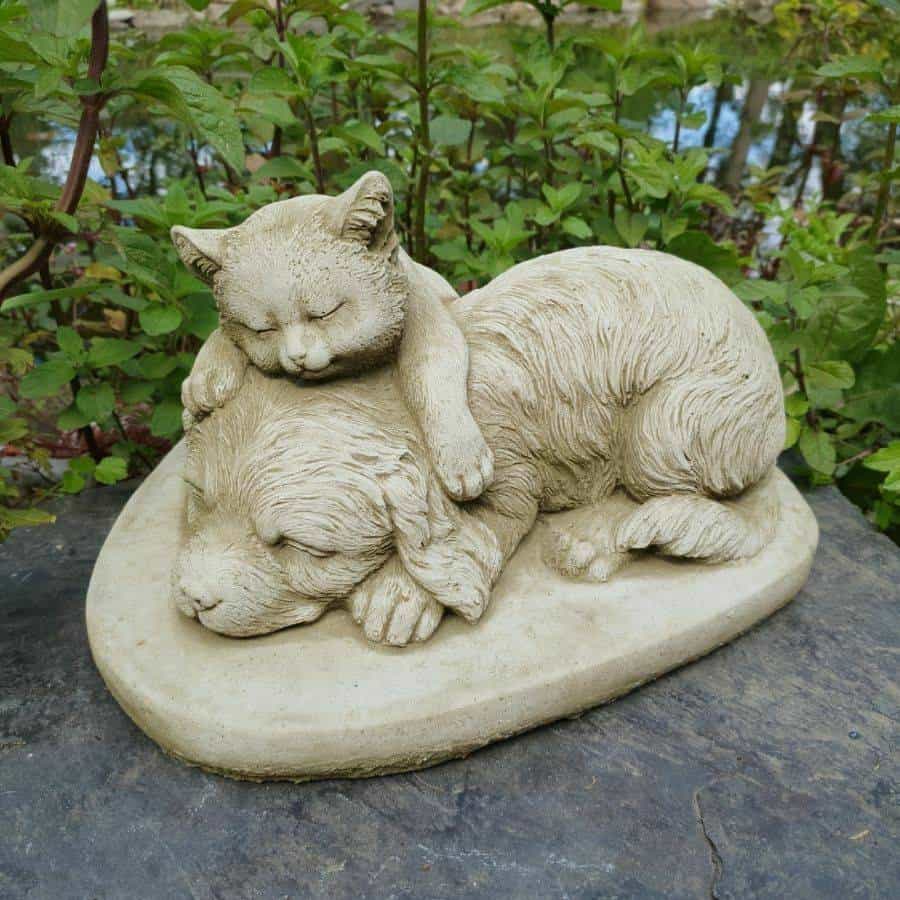 A kitten sleeping and laying on top of a sleeping puppy. Situated on the stone floor of a British garden with green flora in the background