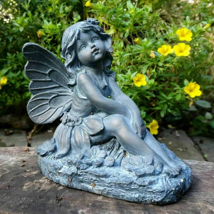 A fairy statue with delicate wings and peaceful expression, sitting on the wooden log of a British garden with yellow flowers in the background