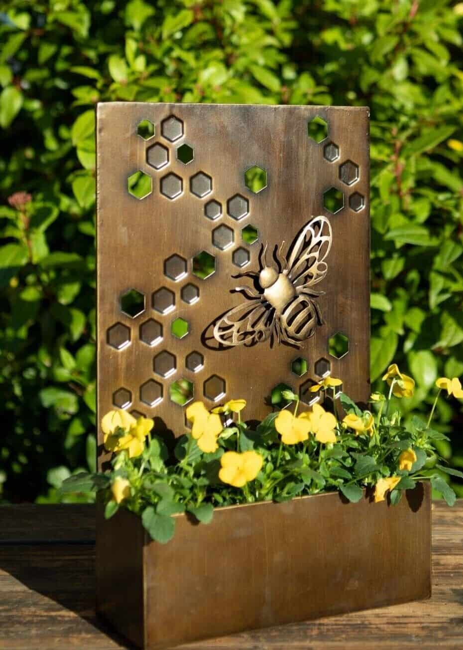 A metal sheet with planter at base with bee design and copper finish. Situated on the decking of a British garden with green foliage in background.