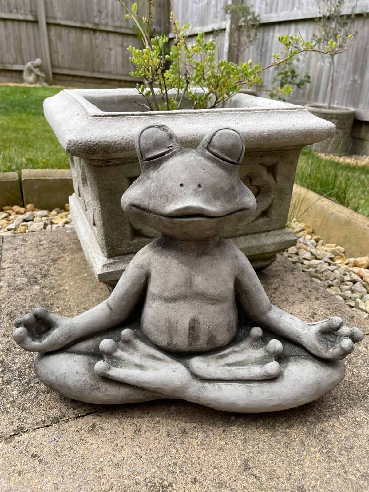 A zen frog sitting with folded legs and extended arms in a yoga position. Situated in the garden of a British home