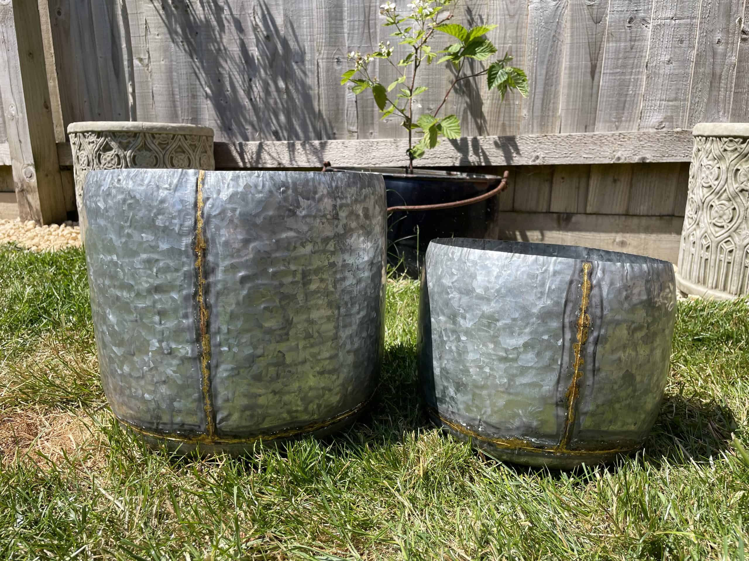 Two plant pots of metal silver finish with golden stripe from top to bottom. Situated in the garden of a British home with green grass and wooden fence