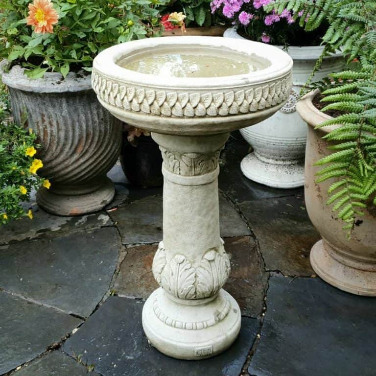 A large birdbath with intricate carvings on base and top rim. Situated in a British garden surrounded by flowerpots