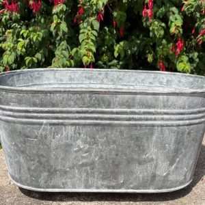 An oval shaped plant pot of galvanised grey metal with hoop handles on the far sides. Situated in a British garden with red flora in the background
