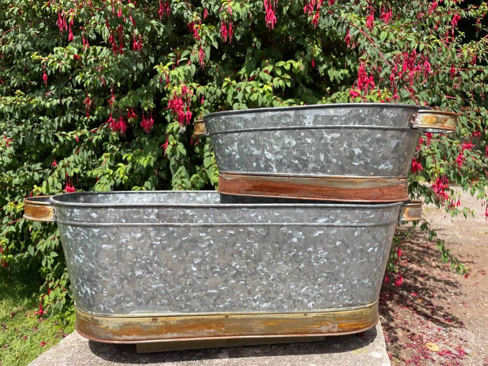 Two planting pots, one large and one smaller sitting on top of the larger. The base of both are wrapped in a copper finish, as are their handles. Situated in a British garden