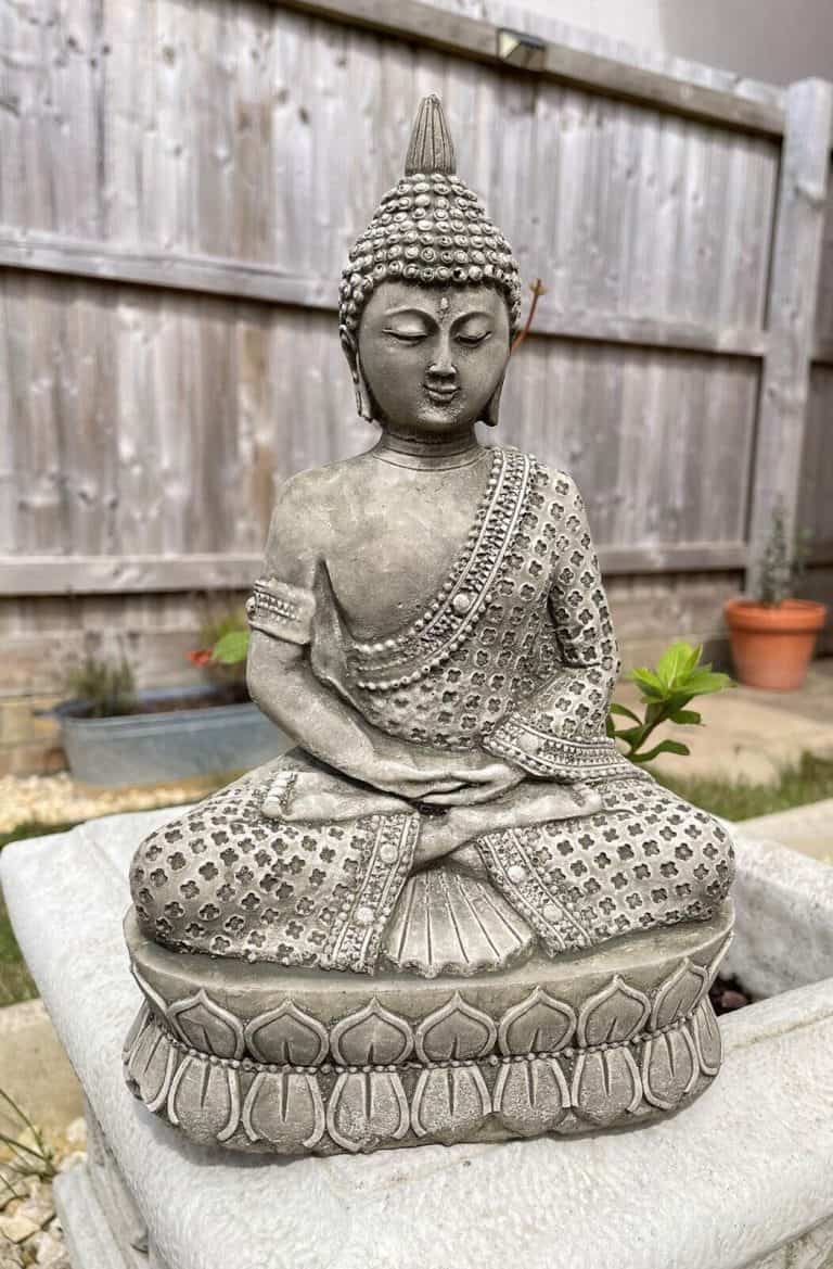 A serene Buddha with regal robes in a meditation pose with arms and legs folded. Situated in a British garden