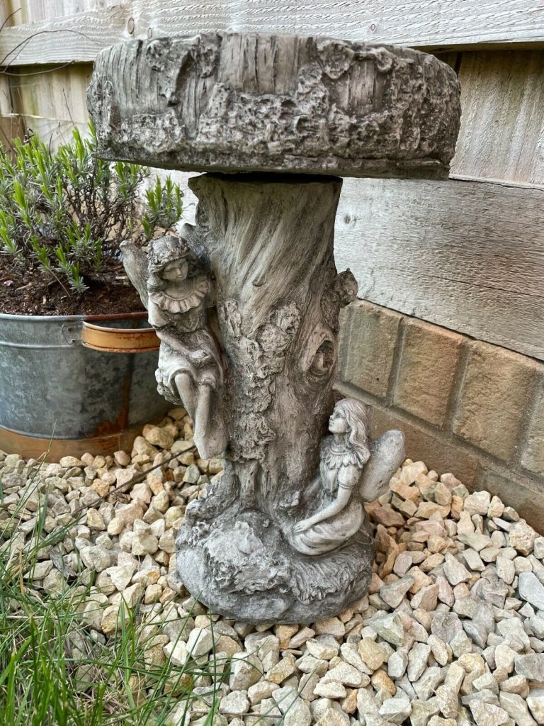 A Beautiful Stone Birdbath with a wooden tree-trunk effect - two lovely female fairies sitting within the branches. Situated in the foreground of a British home's garden