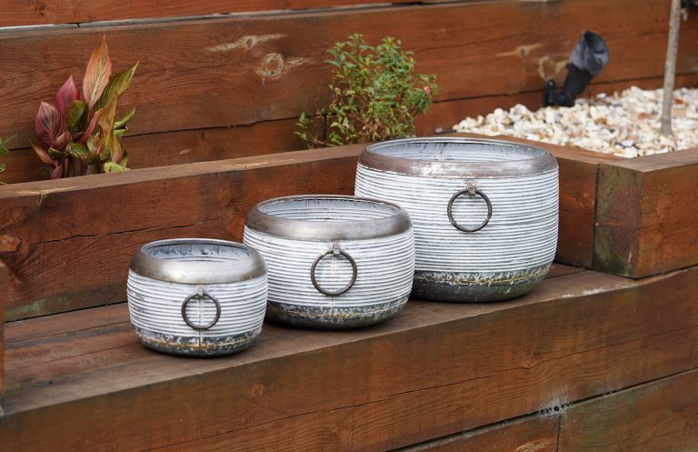 Three Olam planters with white horizontal stipes and bronze handles, base a top. Situated by a brown garden fence in a British Garden