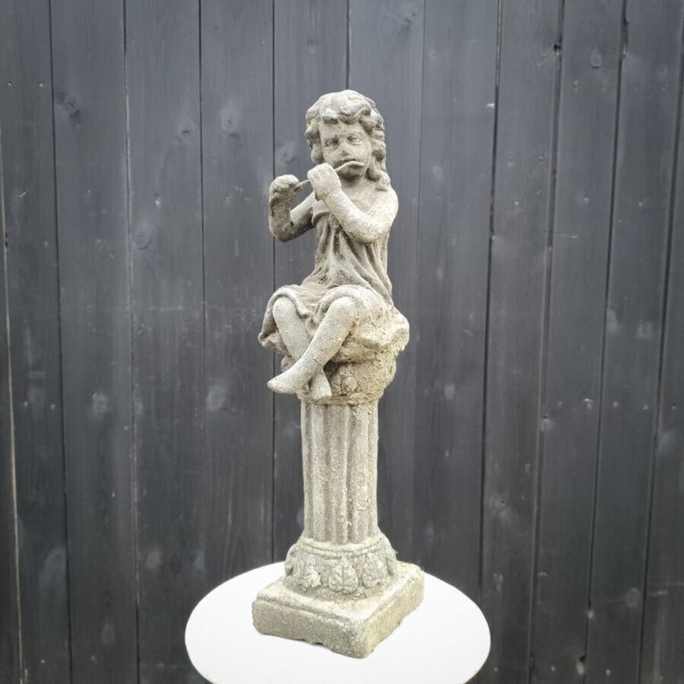 A cherub sitting on a plinth playing a reed. Situated in front of a British garden fence.